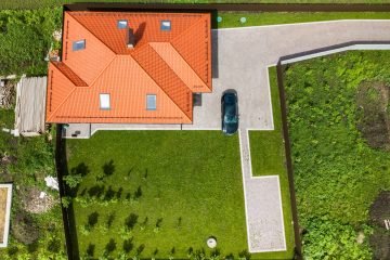 Aerial top view of house shingle roof with attic windows and black car on paved yard with green grass lawn.
